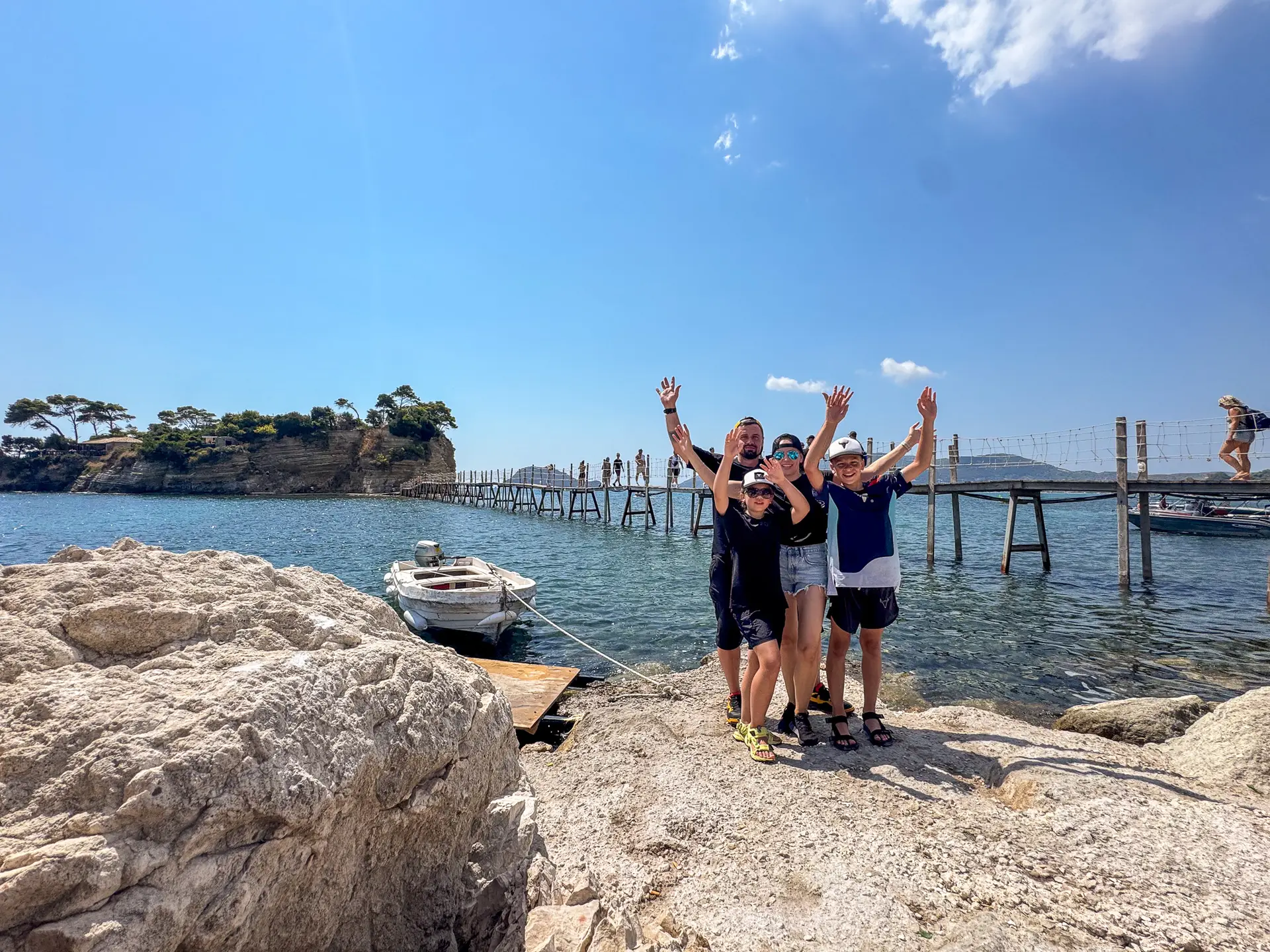 Grupa turystów pozująca z uniesionymi rękami przy drewnianym moście prowadzącym na wysepkę Cameo na Zakynthos.