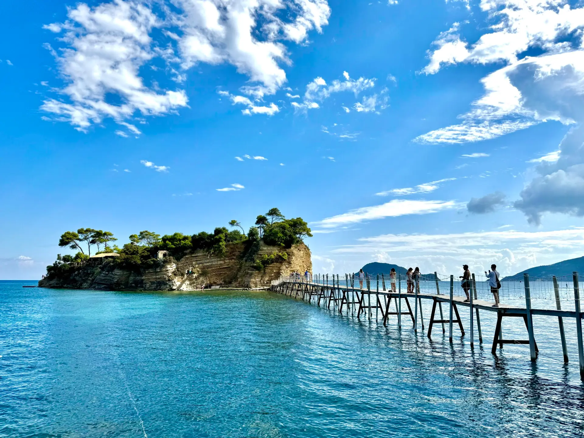 Most prowadzący z portu Agios Sostis na wyspę Cameo na Zakynthos, z turkusowym morzem i błękitnym niebem w tle.
