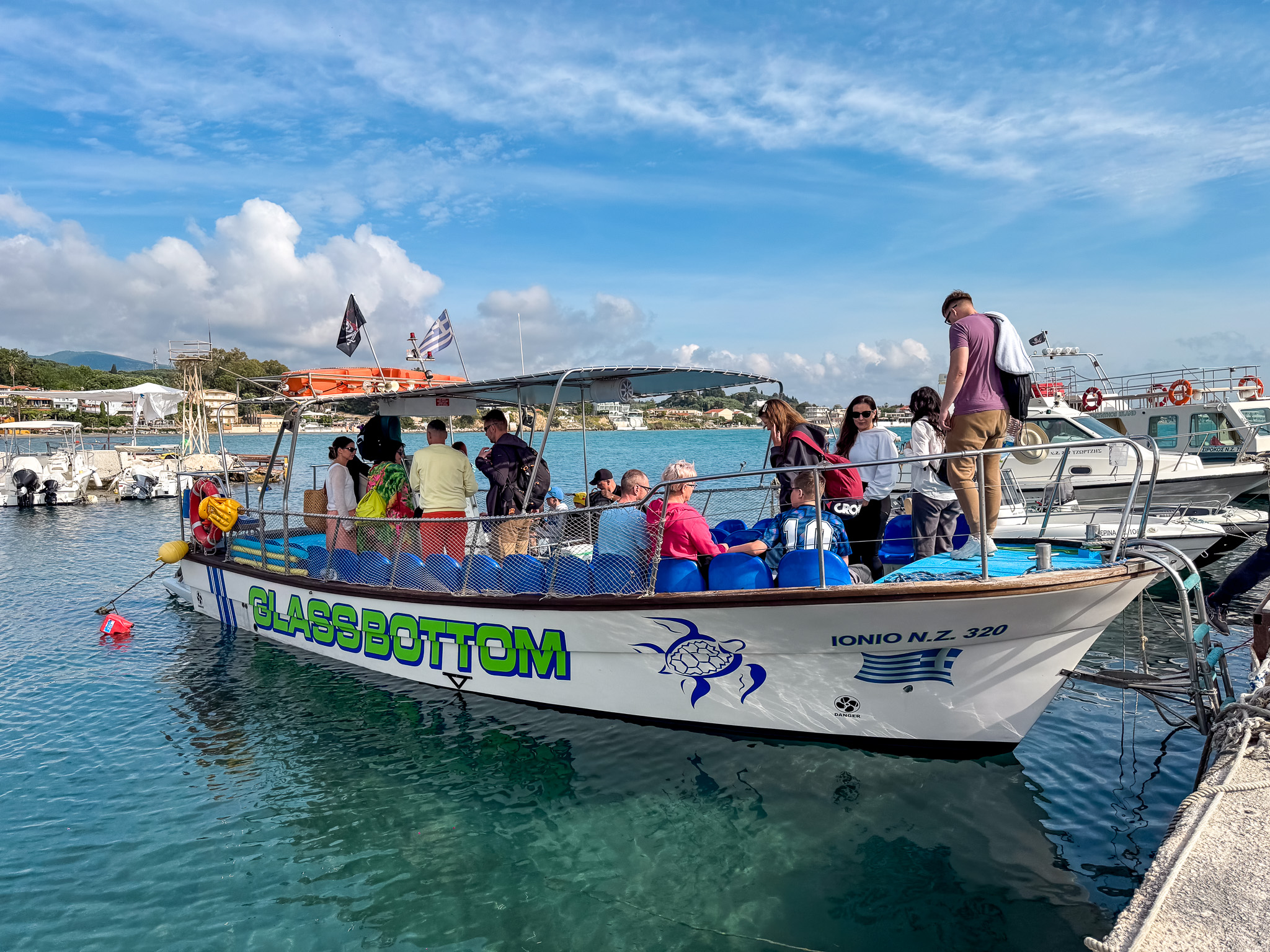 Grupa turystów na łodzi motorowej typu glass bottom z zadaszeniem, przygotowująca się do rejsu po turkusowych wodach Zakynthos.